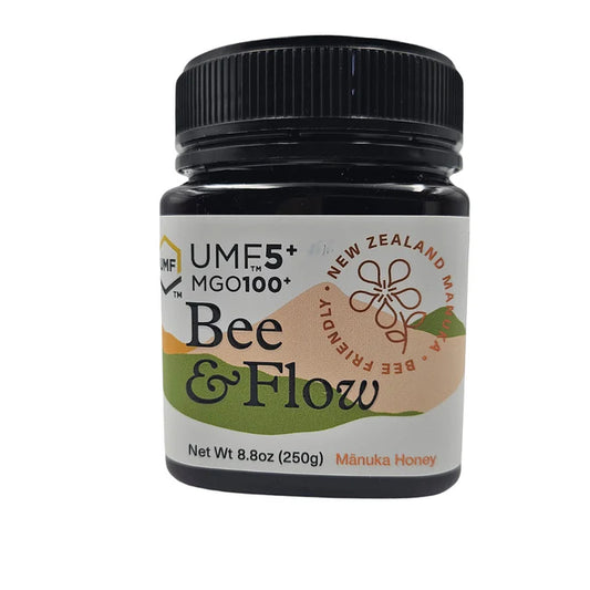 Jar of WellNest Bee & Flow Raw Manuka Honey UMF 5+ with MGO 100+, amber honey with a label and wooden honey dipper.